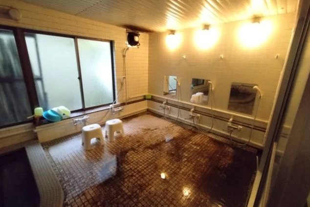 寮の大浴場。複数のシャワー、鏡、広い浴槽、窓が映る共用スペース。