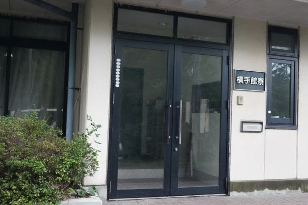 寮の外観。「横手館寮」と書かれた看板と黒いガラス扉の入り口が映る。