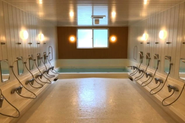 清潔感のある白いタイル張りの空間に、複数のシャワーと鏡が並び、奥には湯船が見える大浴場が写っている画像。リゾートバイトの寮に完備された共同風呂のイメージ。