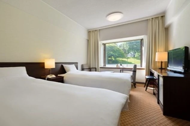 ホテル・旅館の客室。ベッド2台、机、テレビ、窓から緑が見える快適な部屋が映る寮のイメージ画像。