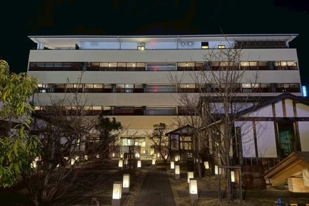 街並みの中に建つ、ベージュとグレーでモダンなデザインのホテル「伊勢神泉」の外観（斜めからのアングル）。上部に切妻屋根の意匠が見える。