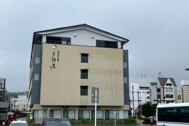 街並みの中に建つ、ベージュとグレーでモダンなデザインのホテル「伊勢神泉」の外観（斜めからのアングル）。上部に切妻屋根の意匠が見える。