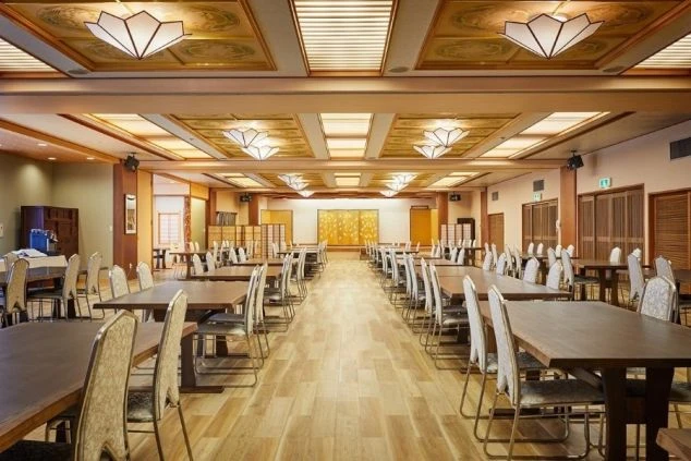 佐渡島のホテルのダイニングの画像。天井には和風の豪華な照明が複数設置されており、部屋全体に木の温かみが感じられる。清潔感のあるテーブルと椅子が規則正しく並べられている。