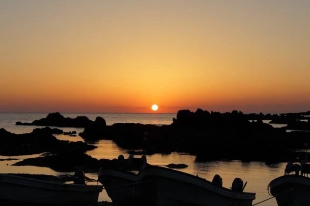 佐渡島の夕焼けの風景の画像。岩場の影から見える夕日が海面に反射しており、手前には漁船のシルエットが写っている。リゾートバイトの休日に観光を楽しんでいる風景の画像。