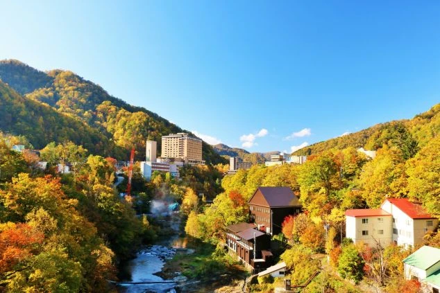 紅葉に染まる山々に囲まれた温泉街の風景の画像。川沿いには旅館やホテルが建ち並んでおり、秋の美しい景色が広がっている。リゾートバイトの風景の画像。