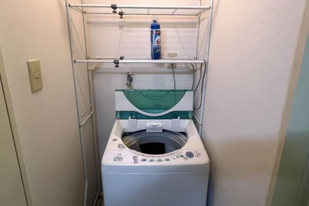 リゾートバイトの寮に備え付けられた洗濯機の画像。棚には洗剤が置かれ、縦型の洗濯機が設置されている。リゾートバイトの寮の設備の画像。
