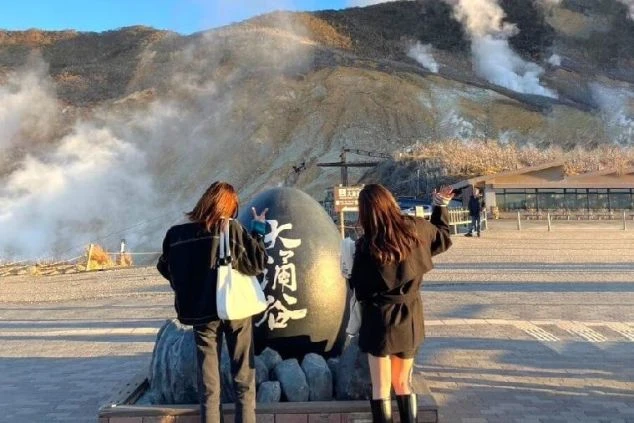大涌谷の噴煙を背景に、2人の女性が記念撮影している後ろ姿の画像。リゾートバイトの休日に温泉地の絶景を楽しむイメージ。