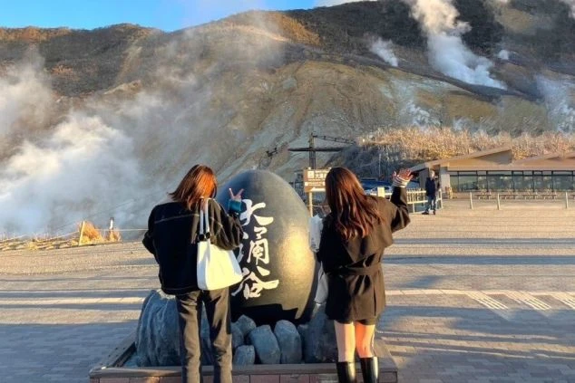 大涌谷の噴煙を背景に、2人の女性が記念撮影している後ろ姿の画像。リゾートバイトの休日に温泉地の絶景を楽しむイメージ。