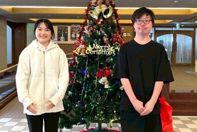 クリスマスツリーを挟んで並ぶ、白いパーカーの女性と黒いTシャツの男性。ホテルのロビーのような場所で笑顔を見せている。