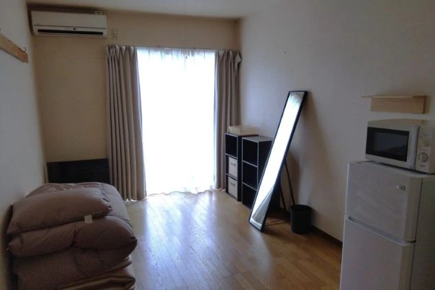 リゾートバイトの寮の部屋の画像。フローリングの床に、布団と姿見、収納ラックが置かれている。窓からは明るい光が差し込み、エアコンが備え付けられている。リゾートバイトの寮の部屋の画像。