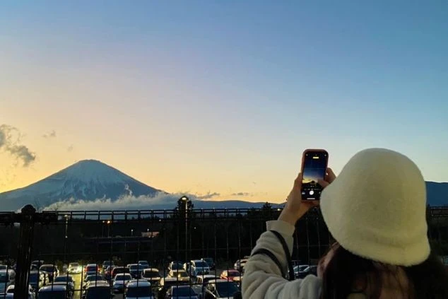 夕焼けに染まる雄大な富士山を、スマートフォンで撮影する女性の画像。