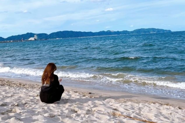 リゾートバイトの休日の過ごし方。ビーチで海を眺めながら座っている女性の後ろ姿。