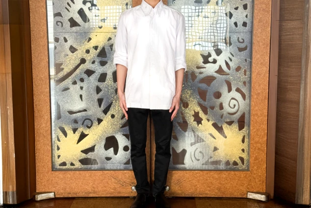 厨房の制服を着た男性。白い七分袖のコックコートと黒いズボンを着用している。