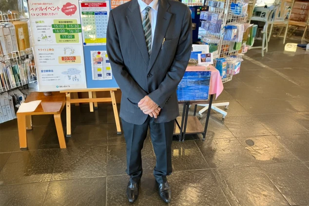  フロントスタッフの制服を着た男性。グレーのスーツとストライプのネクタイを着用し、ロビーの掲示板の前に立っている。