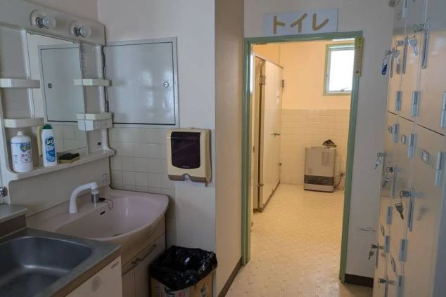 リゾートバイトの寮の共用スペースにある洗面台の画像。洗面台の横にはゴミ箱があり、奥の扉の向こうにはトイレが見える。共用スペースの洗面台と、ロッカーが並んでいる。リゾートバイトの寮の共用スペースの画像。