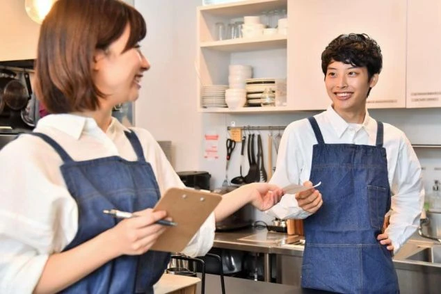 2人の女性スタッフが笑顔で接客している画像。一人はバインダーを持ち、もう一人は伝票を受け取っている。リゾートバイトの仕事内容の一つである、レストランホールスタッフの仕事風景の画像。