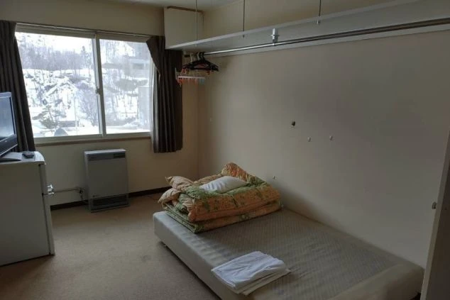 リゾートバイトの寮の部屋の画像。部屋にはベッドと小さなテレビ、冷蔵庫が置かれている。窓の外には雪景色が見え、ハンガーラックも備え付けられている。リゾートバイトの寮の部屋の画像。