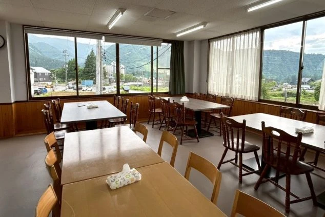 リゾートバイトの寮の食堂。窓から山々が見え、広々とした空間にテーブルと椅子が多数並んでいる。賄いを食べる場所として、開放的な雰囲気を伝える。