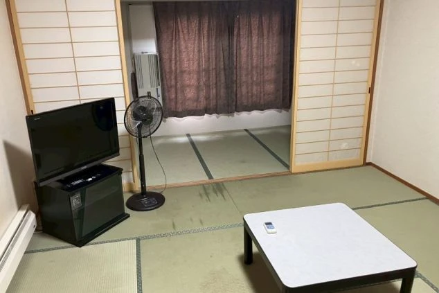 リゾートバイトの寮の和室。テレビ、扇風機、テーブルが備え付けられており、奥の部屋にはエアコンも見える。畳の部屋でくつろげる雰囲気をアピール。