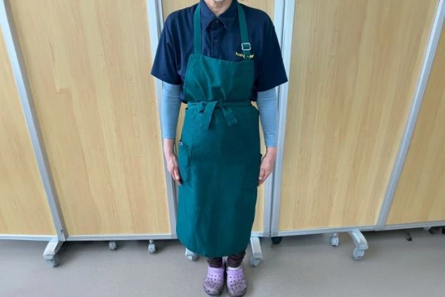 紺色のポロシャツと緑色のエプロンを着用した男性スタッフの画像。ポロシャツの胸には刺繍のロゴが入っている。リゾートバイトの仕事内容を表す、従業員食堂スタッフの制服の画像。