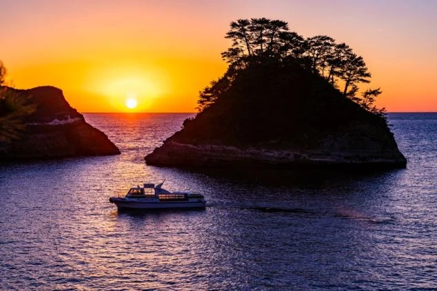 夕焼けに染まる西伊豆の堂ヶ島の画像。海に浮かぶ島々を背景に、遊覧船が通る様子を捉えた写真。