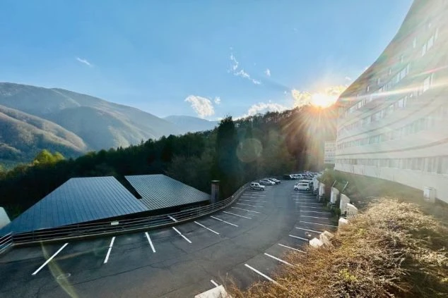 日が差す山間のリゾートホテルの外観の画像。なだらかなカーブを描いた建物が、駐車場の周りの緑に囲まれている。リゾートバイトの仕事場となる、ホテルの外観の画像。