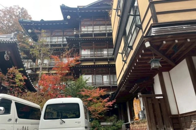 外観。左奥には千と千尋の神隠しを彷彿とさせる、複雑な構造と瓦屋根を持つ伝統的な木造建築の建物がそびえ立ち、手前には比較的新しい黄土色の棟が並んでいる。手前には紅葉した木々や白い軽トラックが見える。