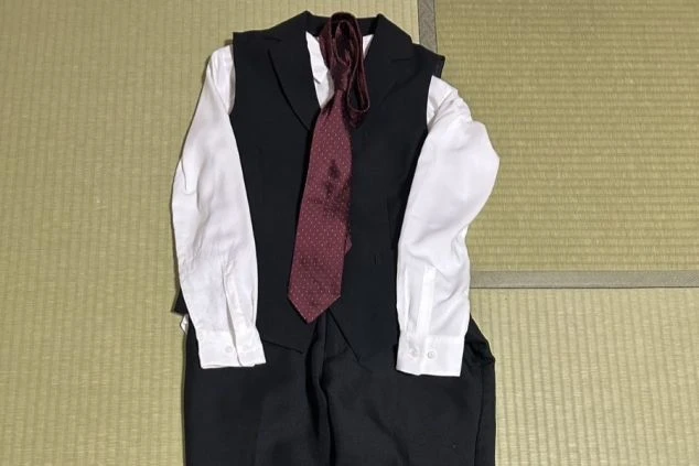 畳の上に置かれた、レストランスタッフの制服画像。白い長袖シャツに黒いベストとパンツ、ワインレッドのドット柄のネクタイのセット。