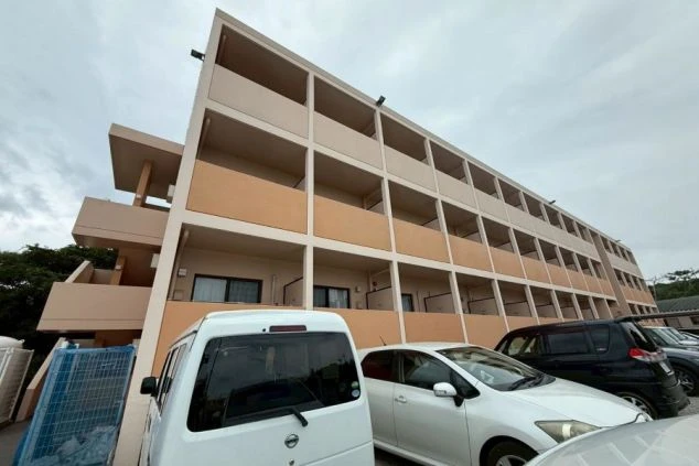 寮の外観。ベージュとオレンジの壁で、バルコニーが格子状に並ぶ多層階の建物。手前には駐車スペースと複数の車が見える。