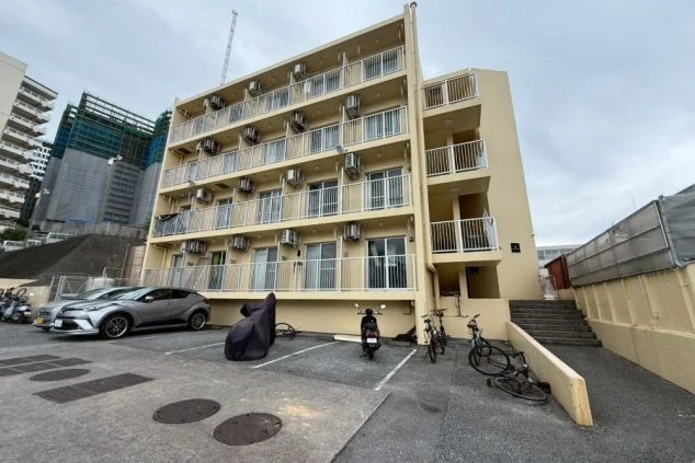 寮の外観。薄いベージュ色の5階建ての建物で、各階にエアコンの室外機とバルコニーが並んでいる。手前は駐車場で、数台の車やバイクが停まっている。