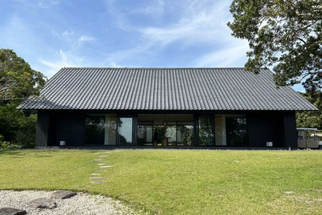 建物の正面。大きな瓦屋根と黒い壁のモダンな和風建築で、全面ガラス戸の向こうに室内が見える。手前は手入れされた芝生で、飛び石がある。