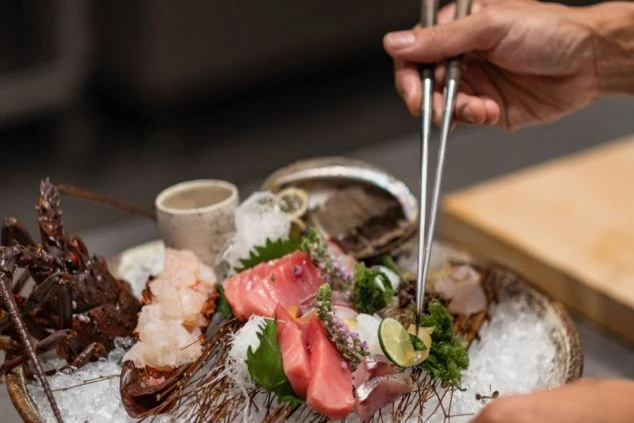 建物の正面。大きな瓦屋根と黒い壁のモダンな和風建築で、全面ガラス戸の向こうに室内が見える。手前は手入れされた芝生で、飛び石がある。