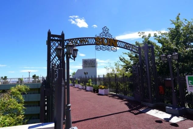 リゾートバイトの勤務先の外観を表現するイメージ画像。青空の下に「SUN」という文字が飾られたアイアンゲートがあり、その先には海へと続く遊歩道が広がっている。