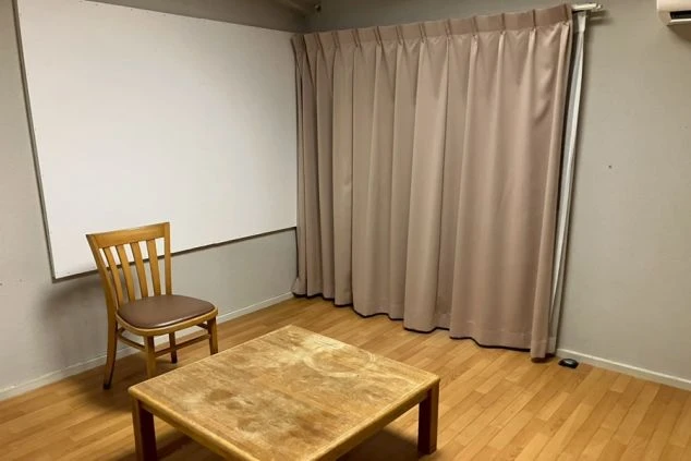 リゾートバイトの寮の個室。木製のテーブルと椅子、カーテンが備え付けられている、シンプルな内装の部屋です。