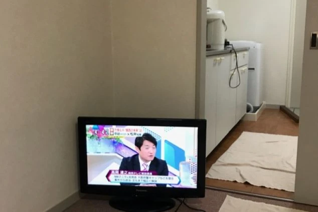 寮の部屋の画像。テレビが置かれており、奥にはキッチンや洗濯機が見える。