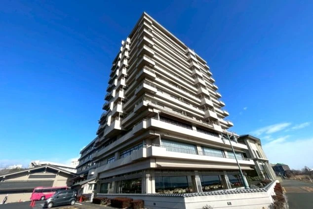 琵琶湖グランドホテルの正面外観。低層のロビー棟と、段々になったバルコニーが特徴的な高層棟。手前はアスファルトの駐車場で、奥には琵琶湖が見える。