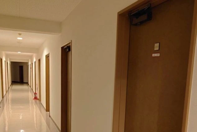 寮の長い廊下。両側に居室のドアが並び、奥まで明るく照らされている。
