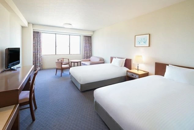 2つのベッドが並べられた、広々としたホテルの客室の画像。窓の外には景色が広がり、部屋にはソファーやテレビ、デスクが備え付けられている。リゾートバイトの寮やホテルの客室の画像。