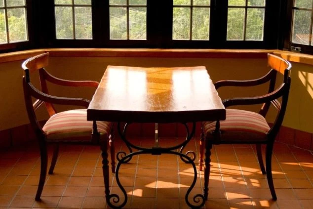 タイル張りの床に、テーブルと椅子が2つ置かれた明るいテラスの画像。大きな窓から光が差し込み、外の緑が見える。