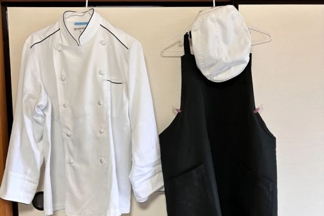 ハンガーにかけられた、調理スタッフの制服の画像。白いコックコートと黒いエプロン、白い帽子がセットになっている。清潔感のある制服で、リゾートバイトの仕事内容を表す画像の画像。