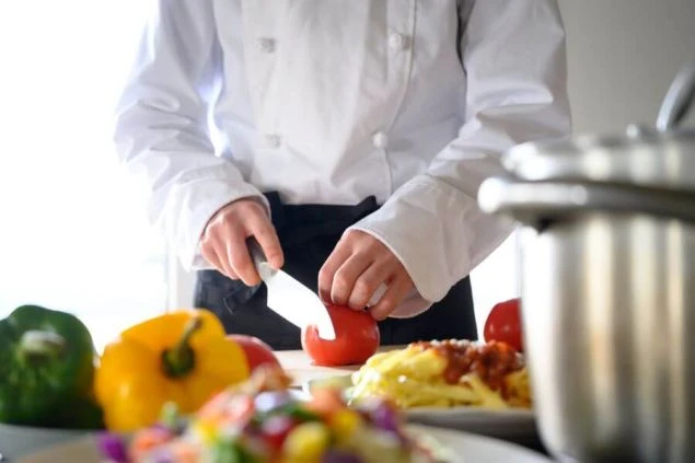 シェフの制服を着用したスタッフが、料理を調理している画像。まな板の上のトマトを包丁でカットしている。リゾートバイトの仕事内容を表す、調理スタッフの仕事風景の画像。