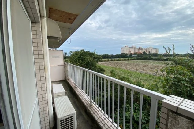 寮のバルコニー。エアコン室外機と広い野原、大きな建物が映る。