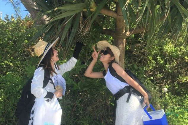 沖縄リゾートバイトを楽しむ笑顔の女性2人。麦わら帽子をかぶり、白い衣装で南国フルーツらしきものを指差している。日差しが降り注ぐ中、リゾート地での仕事と休日を満喫するイメージ。