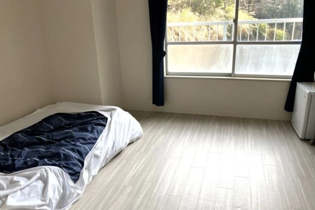 ワクトリの社員が出張で撮影した、リゾートバイトの寮の部屋の画像。フローリングの広々とした空間に、布団と小型冷蔵庫が置かれ、窓からは明るい日差しが差し込んでいる。