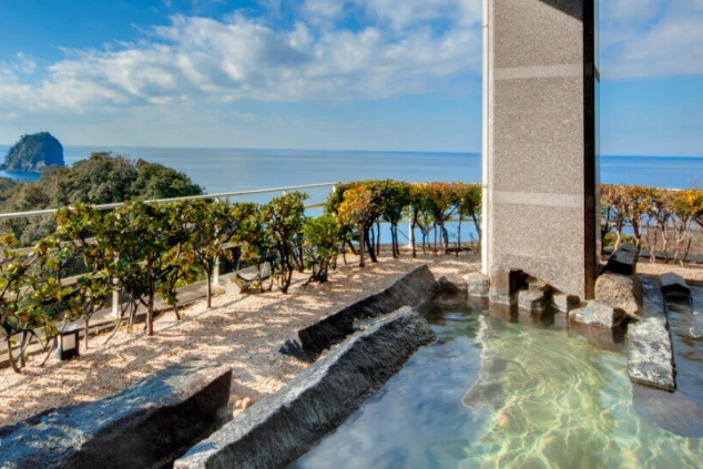 リゾートホテルの露天風呂。大きな岩風呂の向こうに、青い海と空、そして岩山が見える。温泉付きのリゾートバイトの魅力を伝える、開放的な雰囲気の画像。