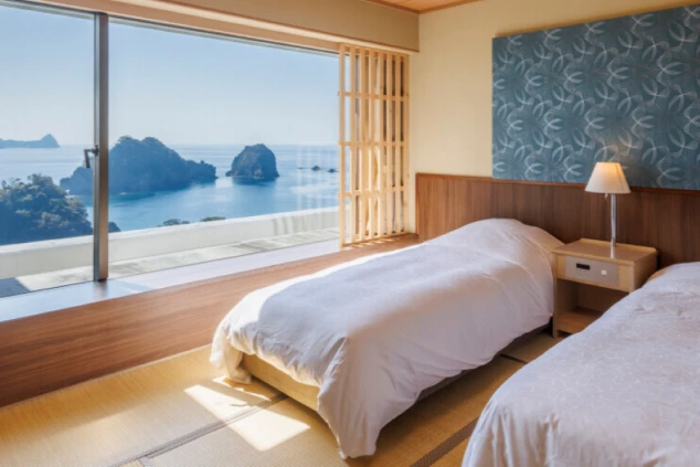 リゾートホテルの和洋室。2つのベッドが置かれ、大きな窓からは穏やかな海と奇岩の絶景が広がる。リゾートバイトの寮や客室の快適さと、素晴らしいロケーションをアピール。