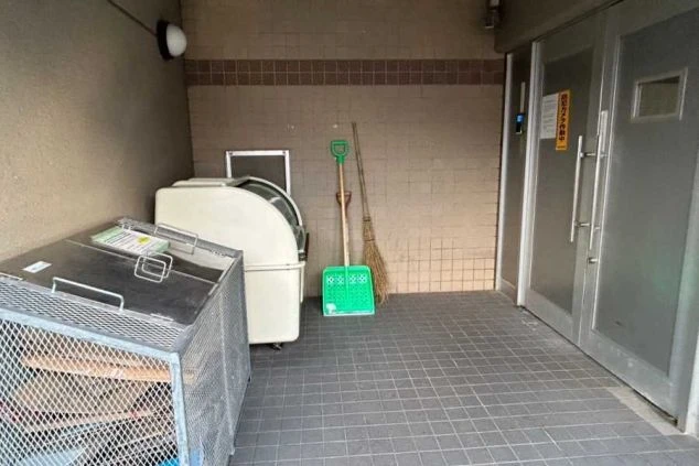 寮のゴミ置き場。屋外用冷蔵庫と清掃用具、防犯カメラ作動中の表示が映る。