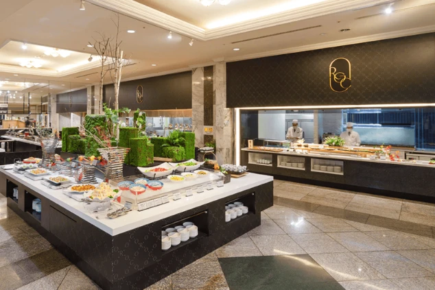  リゾートホテルのレストランにあるビュッフェ。色とりどりの料理が並び、ガラス張りの向こうには調理スタッフの姿が見える。リゾートバイトの仕事場や、食事（賄い）の豪華さをアピールする画像。