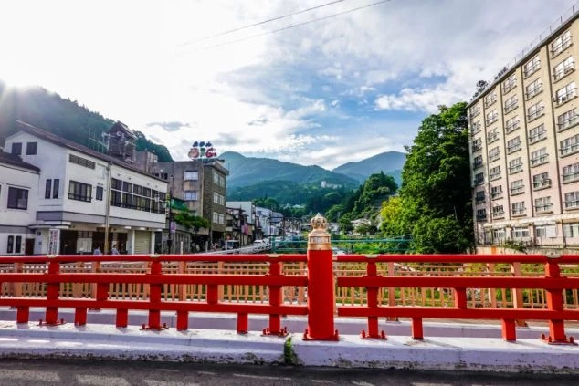 赤い橋と豊かな山々に囲まれた温泉街の風景。川沿いに旅館や建物が並び、自然と共存する街の雰囲気が伝わる。自然豊かな温泉地でのリゾートバイトをアピール。