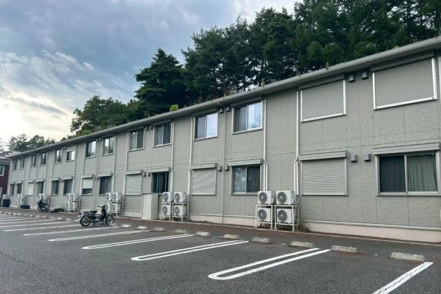 2階建てのリゾートバイトの寮の外観を撮影した画像。部屋の窓にシャッターが付いており、駐車場には車やバイクが停まっている。リゾートバイトの寮の外観の画像。
